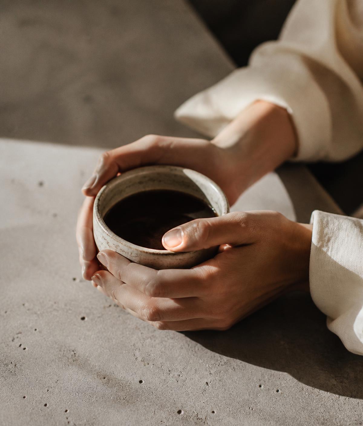 Duas mãos segurando uma xícara de cerâmica com café sobre uma mesa de concreto